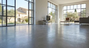 Photographie d'un vaste espace int&eacute;rieur (salon, cuisine ou showroom) avec un sol enti&egrave;rement recouvert d'un rev&ecirc;tement de sol de haute qualit&eacute; (carrelage grand format, b&eacute;ton cir&eacute; liss&eacute; ou r&eacute;sine &eacute;poxy mate) de couleur neutre ou grise, parfaitement fini et homog&egrave;ne, photographi&eacute; depuis un angle qui met en &eacute;vidence la r&eacute;gularit&eacute; et la finition professionnelle du sol, avec un &eacute;clairage naturel mettant en avant la qualit&eacute; de surface et la plan&eacute;it&eacute; du rev&ecirc;tement.
