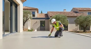Photographie d'un professionnel en équipement de sécurité (gilet jaune, casque) inspectant une surface de sol préparée ou appliquant une chape liquide sur un chantier, mettant en avant la conformité aux normes de sécurité et la qualité de réalisation professionnelle des travaux. L'environnement doit montrer un contexte de chantier professionnel moderne et organisé, illustrant l'engagement de l'entreprise dans la conformité et l'excellence.