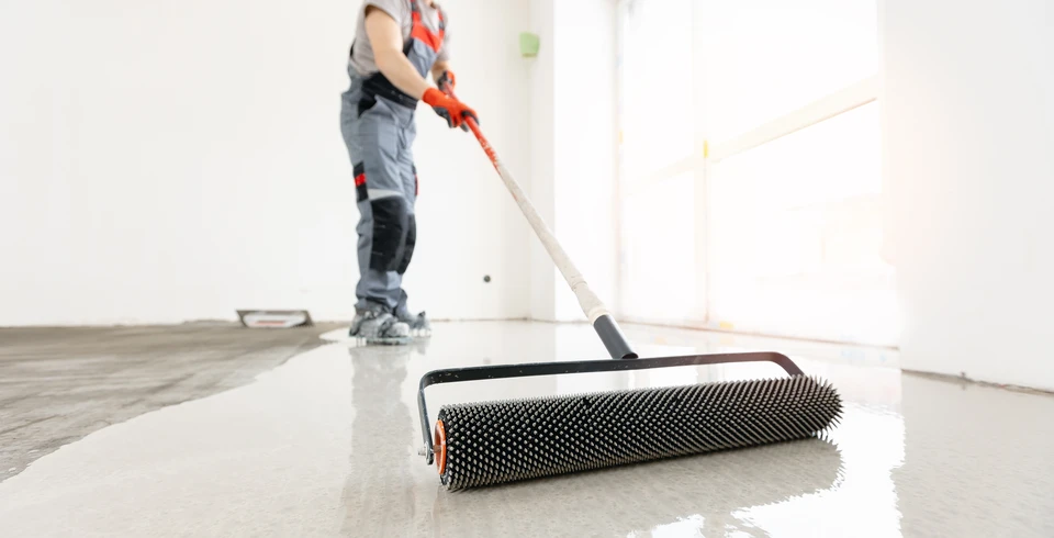 Photographie d'un chantier de travaux de sol en int&eacute;rieur montrant plusieurs &eacute;tapes de finition simultan&eacute;ment&nbsp;: au premier plan, un artisan en tenue professionnelle applique une chape liquide grise sur le sol brut, au centre un deuxi&egrave;me ouvrier pose du carrelage ou un rev&ecirc;tement, et en arri&egrave;re-plan on aper&ccedil;oit des panneaux d'isolation thermique ou de plancher chauffant. L'ambiance doit &ecirc;tre professionnelle, bien &eacute;clair&eacute;e, montrant un chantier structur&eacute; et organis&eacute; avec du mat&eacute;riel de qualit&eacute;. Les ouvriers portent des &eacute;quipements de s&eacute;curit&eacute; appropri&eacute;s (gilets, chaussures de travail). La composition doit transmettre l'id&eacute;e d'une prise en charge globale et coordonn&eacute;e d'un projet complet de sol.