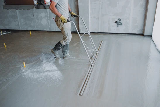Photographie en gros plan d'un artisan posant du parquet ou un rev&ecirc;tement de sol souple en int&eacute;rieur. L'artisan doit &ecirc;tre en &eacute;quipement de travail professionnel (gilet de s&eacute;curit&eacute;, bottes). Il applique le rev&ecirc;tement sur une surface pr&eacute;par&eacute;e en utilisant les outils appropri&eacute;s. On doit voir clairement les lames de parquet ou le sol souple partiellement pos&eacute;, avec les mat&eacute;riaux de finition visibles (colle, joints ou underlay selon le type). L'arri&egrave;re-plan doit montrer un int&eacute;rieur de chantier avec des zones de travail pr&eacute;par&eacute;es. La composition doit mettre en &eacute;vidence le professionnalisme, la technique de pose sp&eacute;cifique au parquet/sol souple et le travail de finition, distinctement diff&eacute;rente des sc&egrave;nes de chape liquide ou de carrelage.