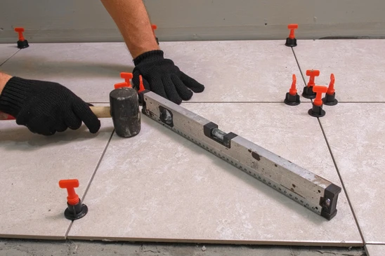 Photographie en gros plan d'un chapiste professionnel en &eacute;quipement de travail (gilet de s&eacute;curit&eacute;, bottes) effectuant le ragr&eacute;age et la pr&eacute;paration d'une surface de sol. L'artisan utilise une taloche ou une spatule de ragr&eacute;age pour lisser et pr&eacute;parer une surface in&eacute;gale, cr&eacute;ant une base uniforme. On voit clairement le compos&eacute; de ragr&eacute;age gris en cours d'application sur une surface en b&eacute;ton brut ou in&eacute;gale, avec le contraste entre les zones pr&eacute;par&eacute;es (lisses) et les zones non encore trait&eacute;es. L'arri&egrave;re-plan montre un chantier int&eacute;rieur avec une structure de b&acirc;timent visible. La composition met en &eacute;vidence le professionnalisme, la technique de pr&eacute;paration pr&eacute;cise et l'importance de cette &eacute;tape fondamentale dans la cha&icirc;ne de rev&ecirc;tement de sol.