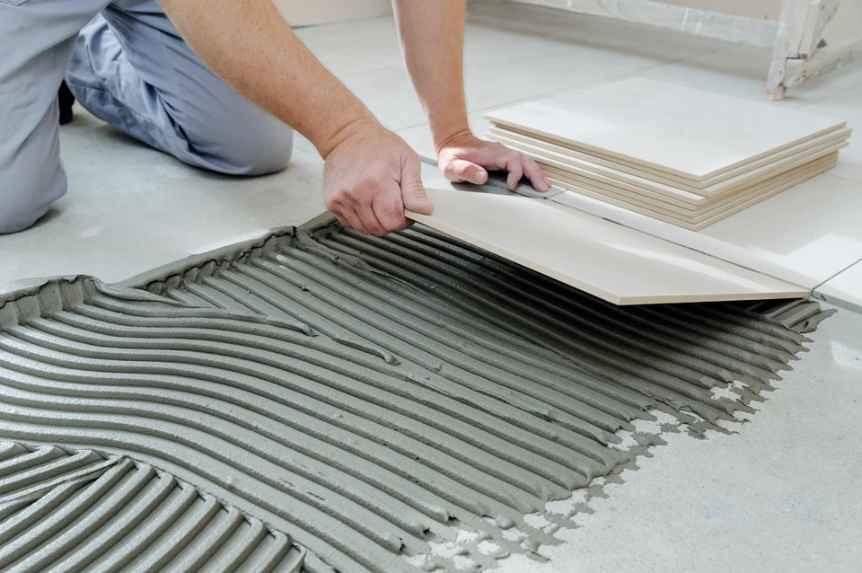 Ouvrier posant carrelage sur sol intérieur, mains gantées appliquant carreaux gris sur mortier-colle