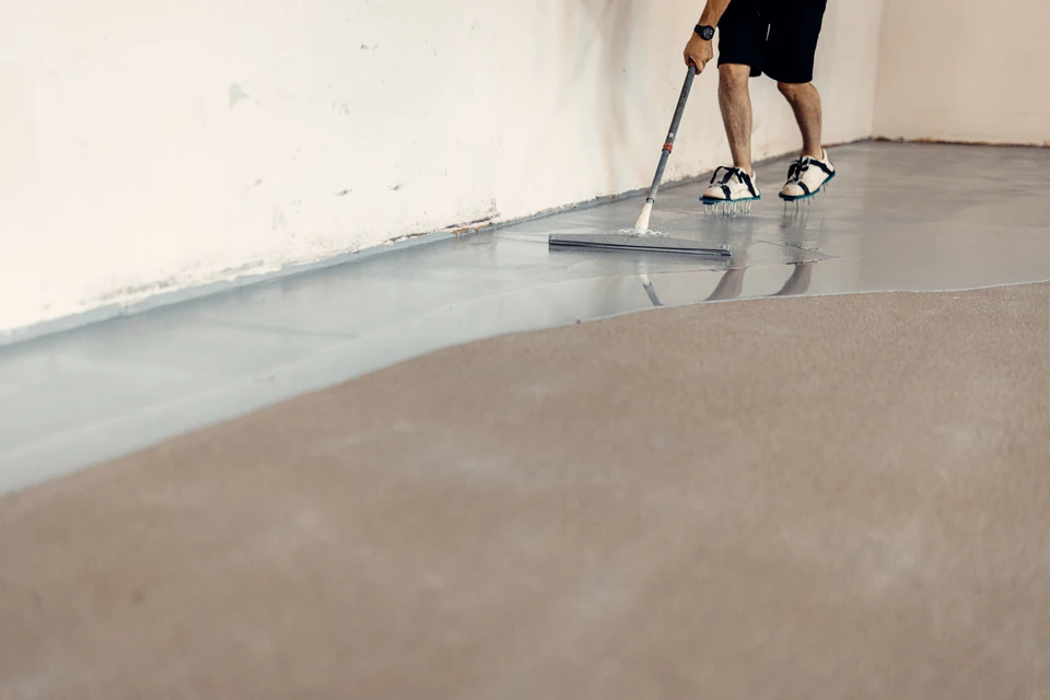 Gros plan sur un artisan en tenue de travail professionnelle appliquant de la résine époxy liquide sur un sol préalablement préparé à l'intérieur d'un bâtiment, utilisant un outil de lissage spécialisé. La surface de travail doit montrer une préparation de qualité (béton ragréé et lisse) et l'artisan concentré sur son travail de finition. La lumière naturelle ou professionnelle doit bien mettre en évidence la texture de la résine en cours d'application et le savoir-faire du professionnel.